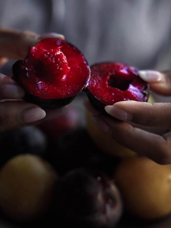 A person gently holds and pulls apart a ripe, juicy red plum, revealing its vibrant, glistening interior. More whole plums and blurred fruit are visible in the background.